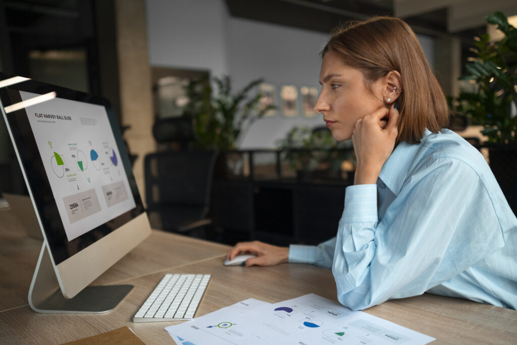 Woman in a light blue shirt rests her chin on her hand while studying charts on a desktop monitor at a wooden desk