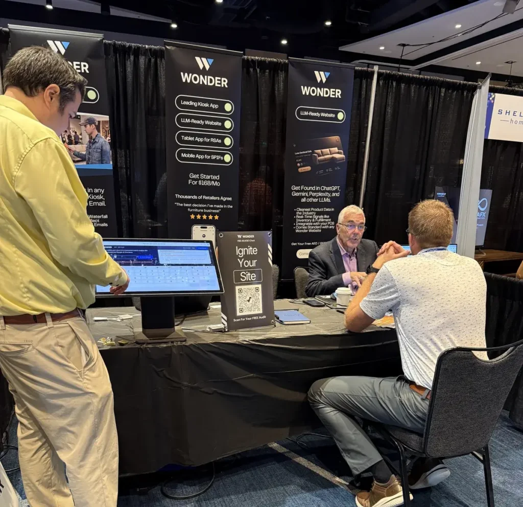 At a trade show booth with three vertical banners that say WONDER; a man in a yellow shirt uses a touchscreen kiosk at a table while two men chat across from him inside a booth.