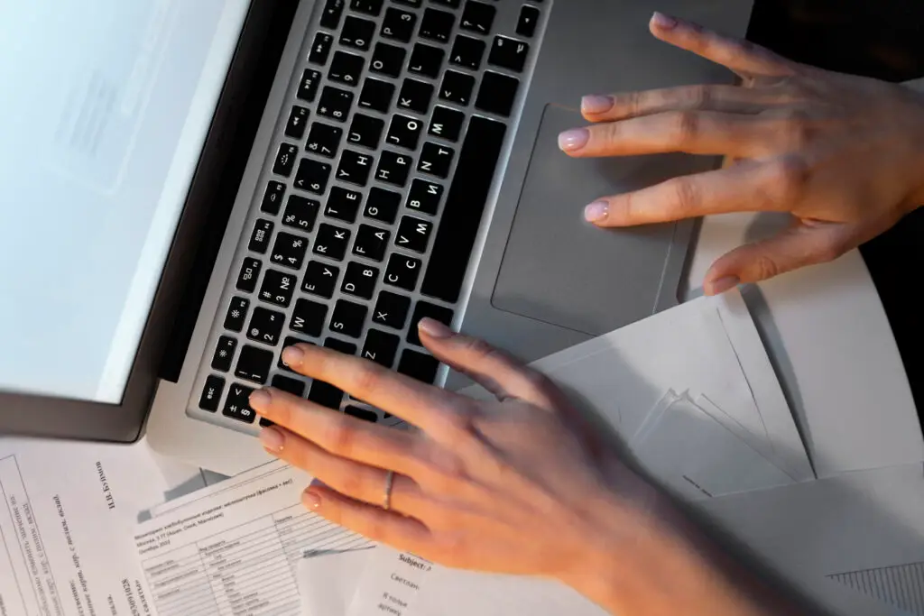 Top down view of two hands typing on a silver laptop keyboard above a desk covered in printed paper documents