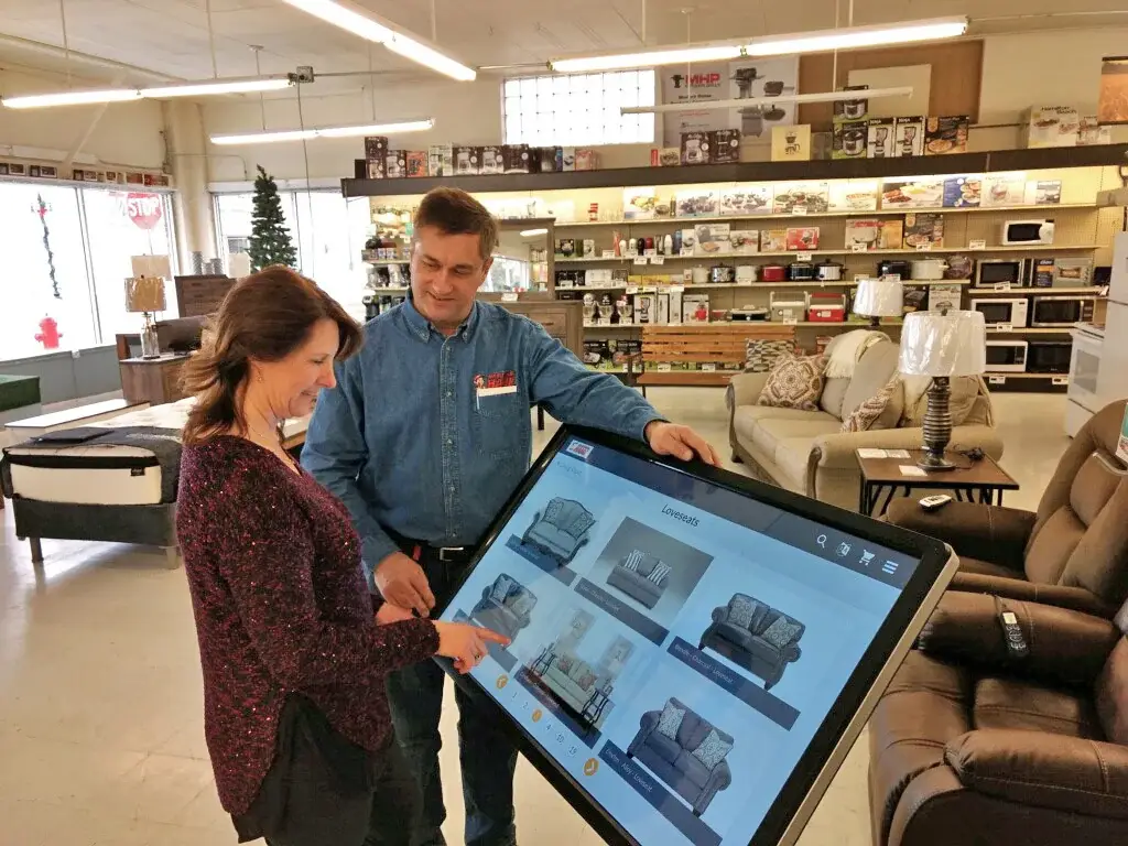Tim Finseth helps a shopper browse loveseats on an endless aisle kiosk beside brown recliners and a sofa Tim Finseth helps a shopper browse loveseats on an endless aisle kiosk beside brown recliners and a sofa