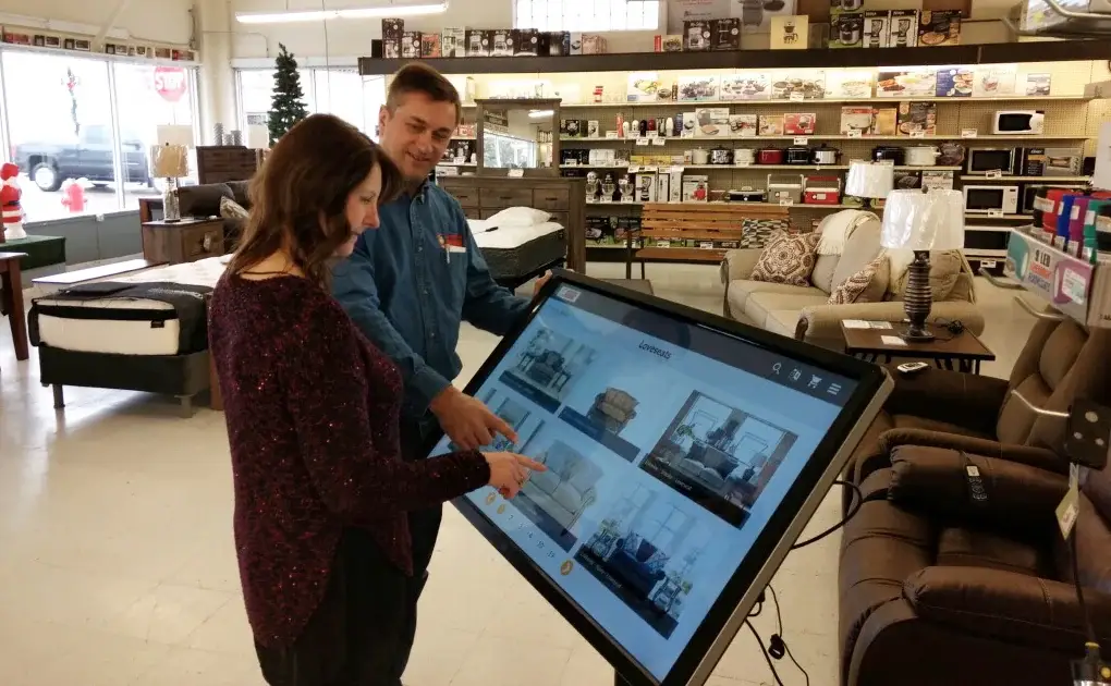 A shopper taps a loveseat on the touchscreen kiosk as Tim Finseth points, with mattresses and lamps behind them A shopper taps a loveseat on the touchscreen kiosk as Tim Finseth points, with mattresses and lamps behind them