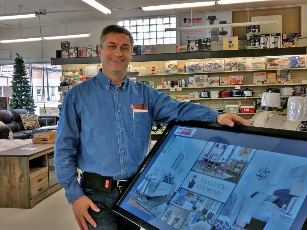 Tim Finseth in a denim Hardware Hank shirt, smiling with one hand on a large touchscreen kiosk in his store Tim Finseth in a denim Hardware Hank shirt, smiling with one hand on a large touchscreen kiosk in his store