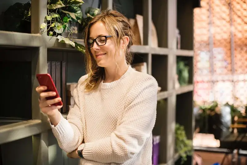 Smiling woman in glasses checking her phone beside a wooden shelving unit at home Smiling woman in glasses checking her phone beside a wooden shelving unit at home