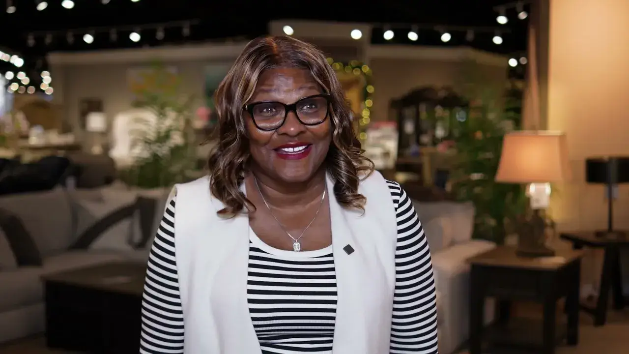 Smiling woman in glasses, a white vest and a striped top stands on a furniture showroom floor with sofas and lamps behind her